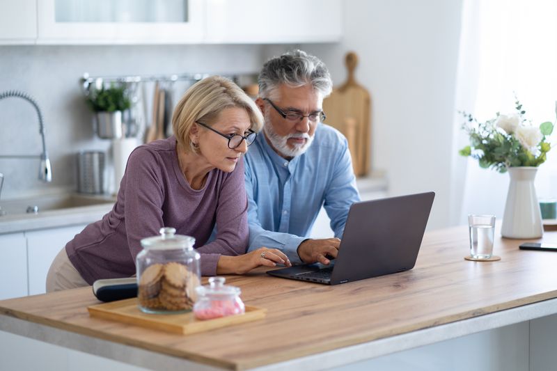 A senior couple review their finances on a laptop in their kitchen. 
