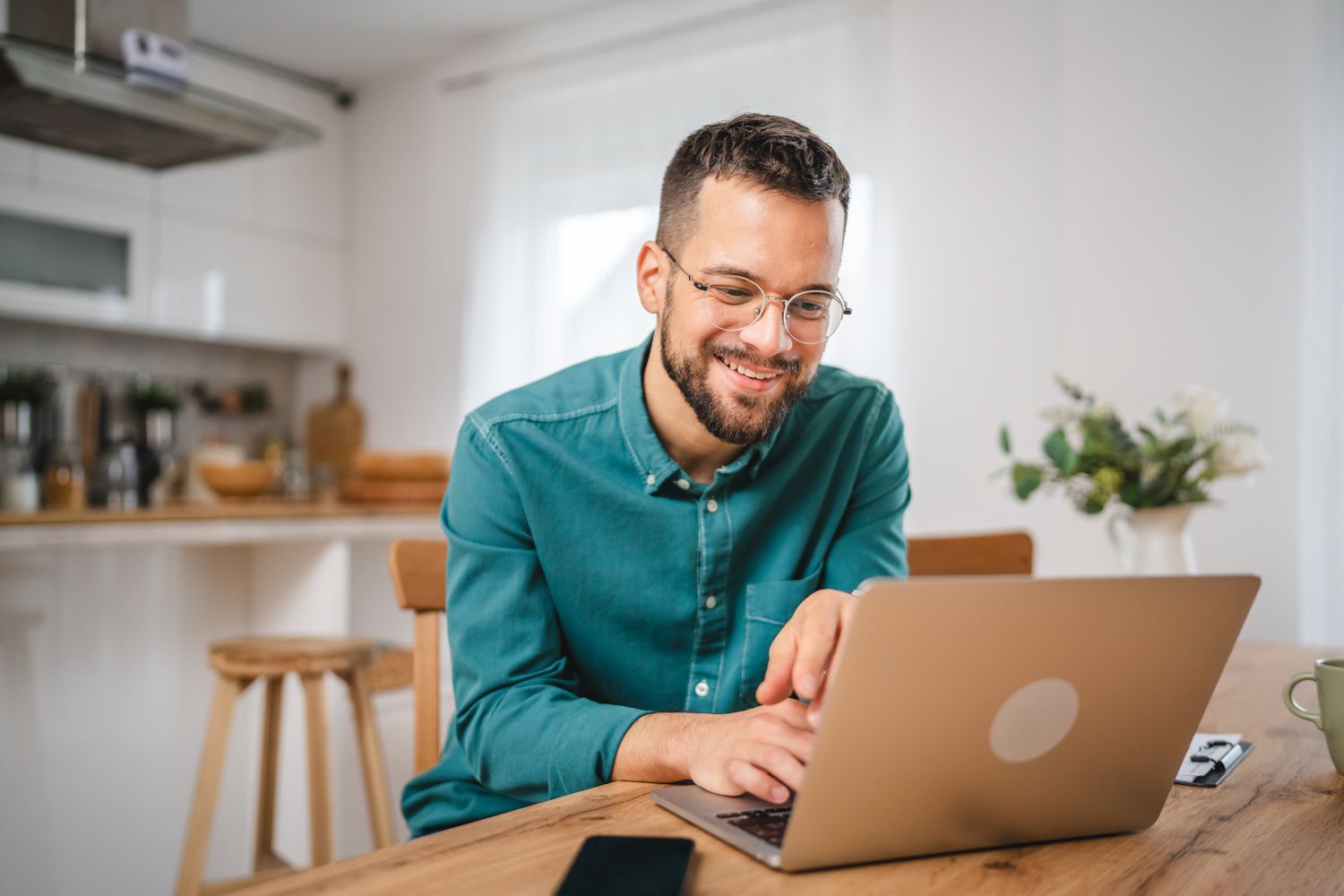 A man sits at a kitchen table, using a laptop and smiling.