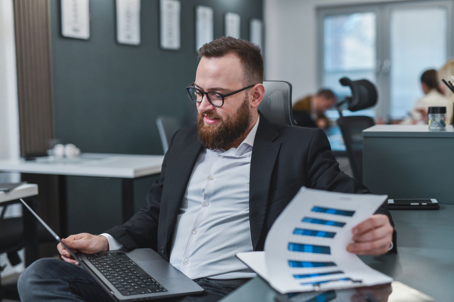 Man in office looking at a laptop while holding document displaying bar charts. 