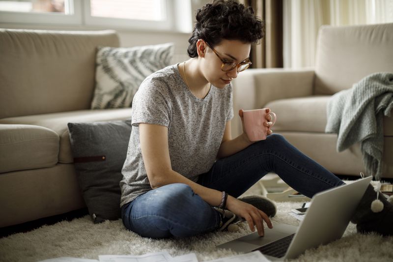 person in grey t-shirt sitting on the floor while drinking coffee and doing work on computer