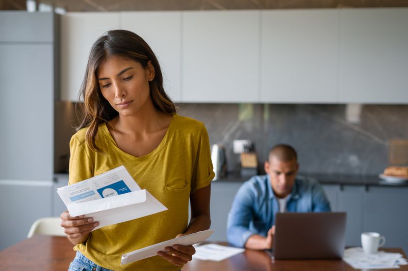 A woman stands in a kitchen looking at mail while a man sits at a table in the back looking at a laptop