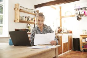 Woman in her 50s sitting at her kitchen table and looking at financial documents and a laptop