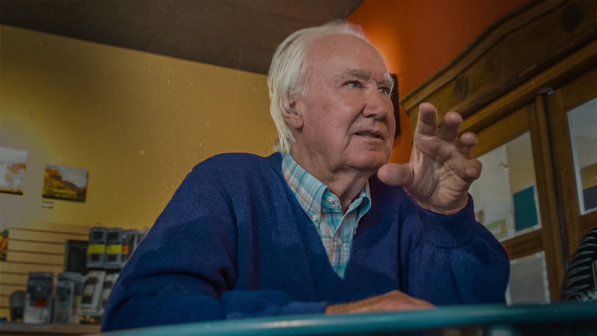 Forrest Fenn gesturing in an indoor setting with shelves and pictures behind him
