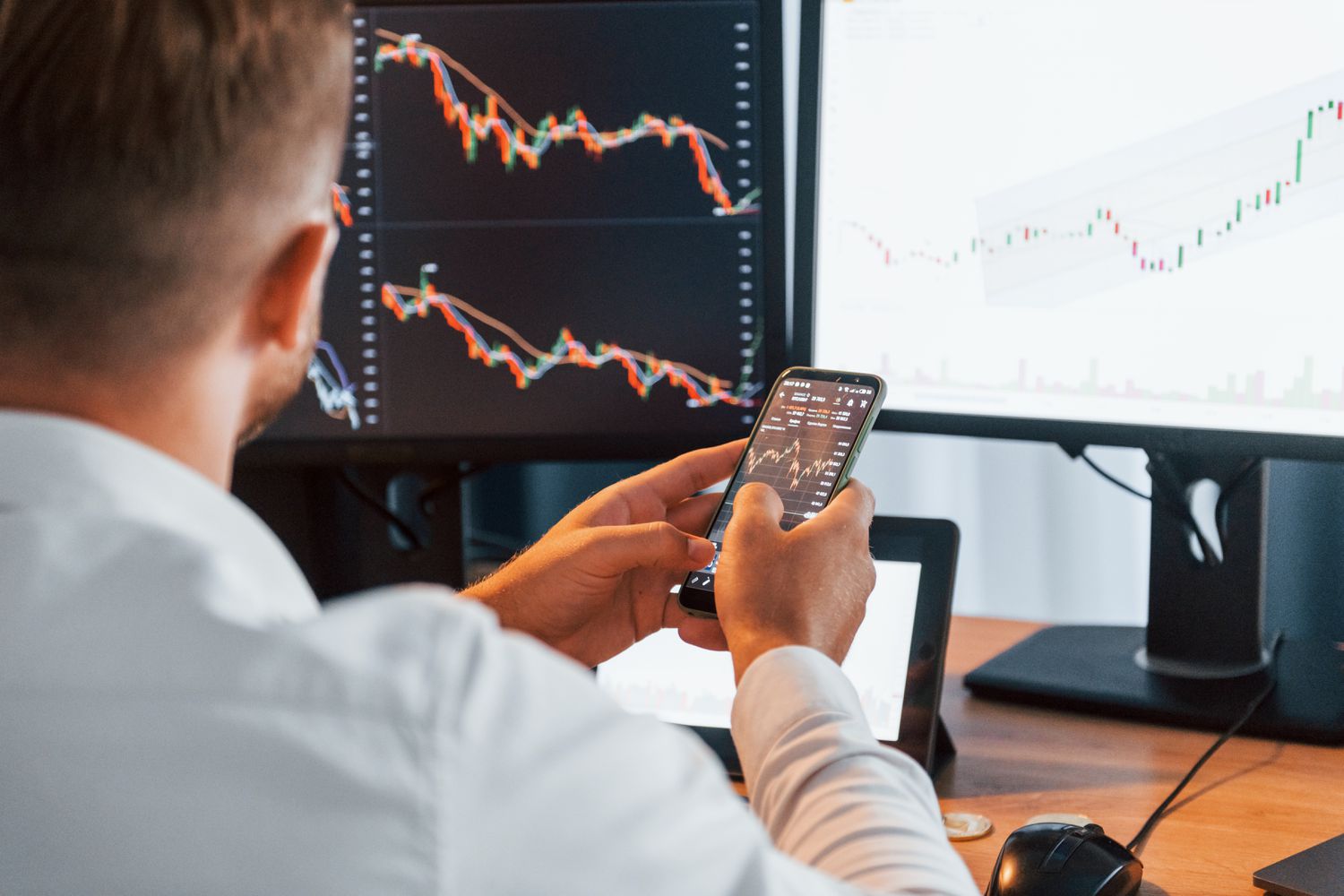 A man with his back to the camera holds a cellphone while looking at two computer screens displaying financial charts.