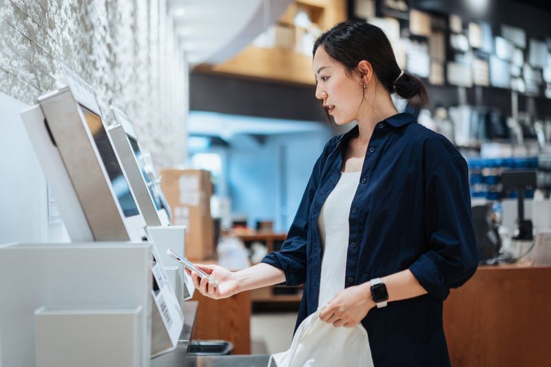 Woman using her phone to checkout a purchase at a self-checkout kiosk.