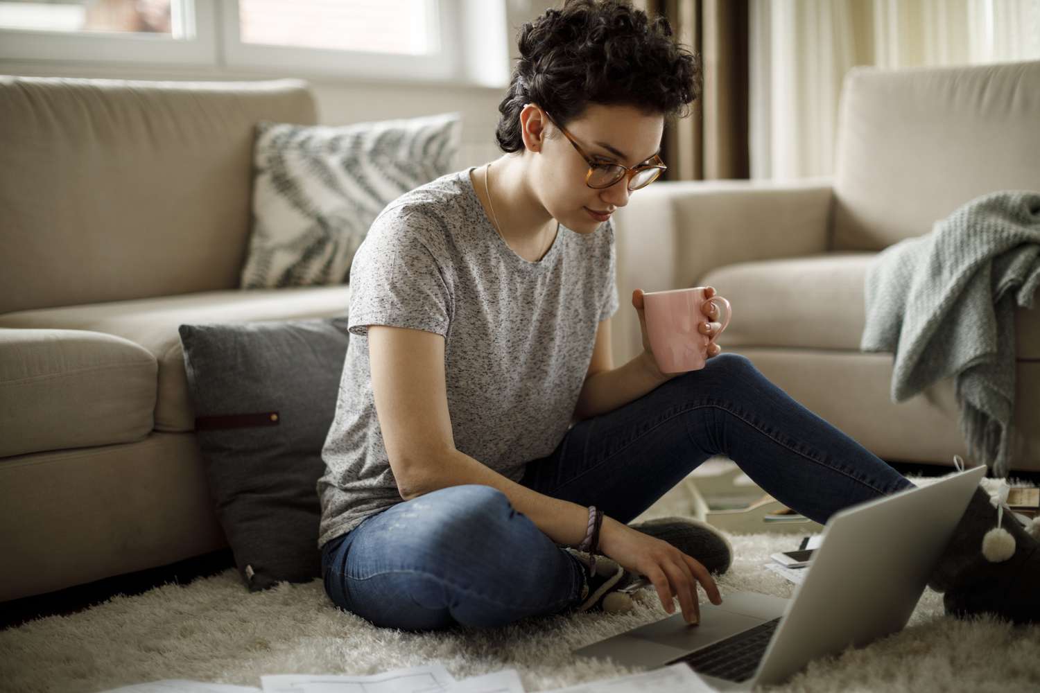 person in grey t-shirt sitting on the floor while drinking coffee and doing work on computer
