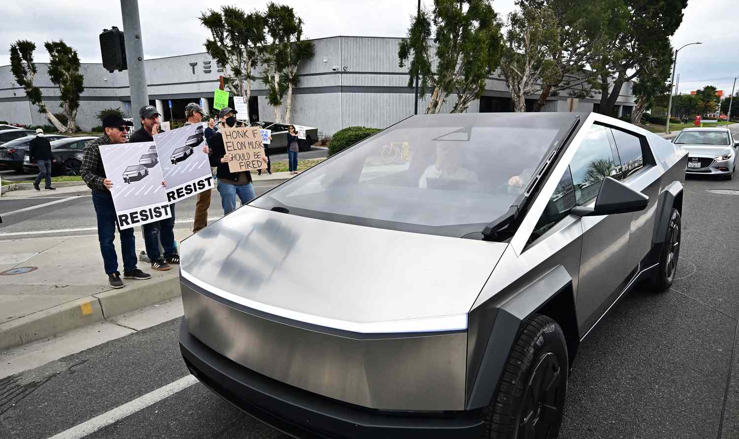Protestors stand between a Tesla dealership and a Tesla Cybertruck driving on the street.