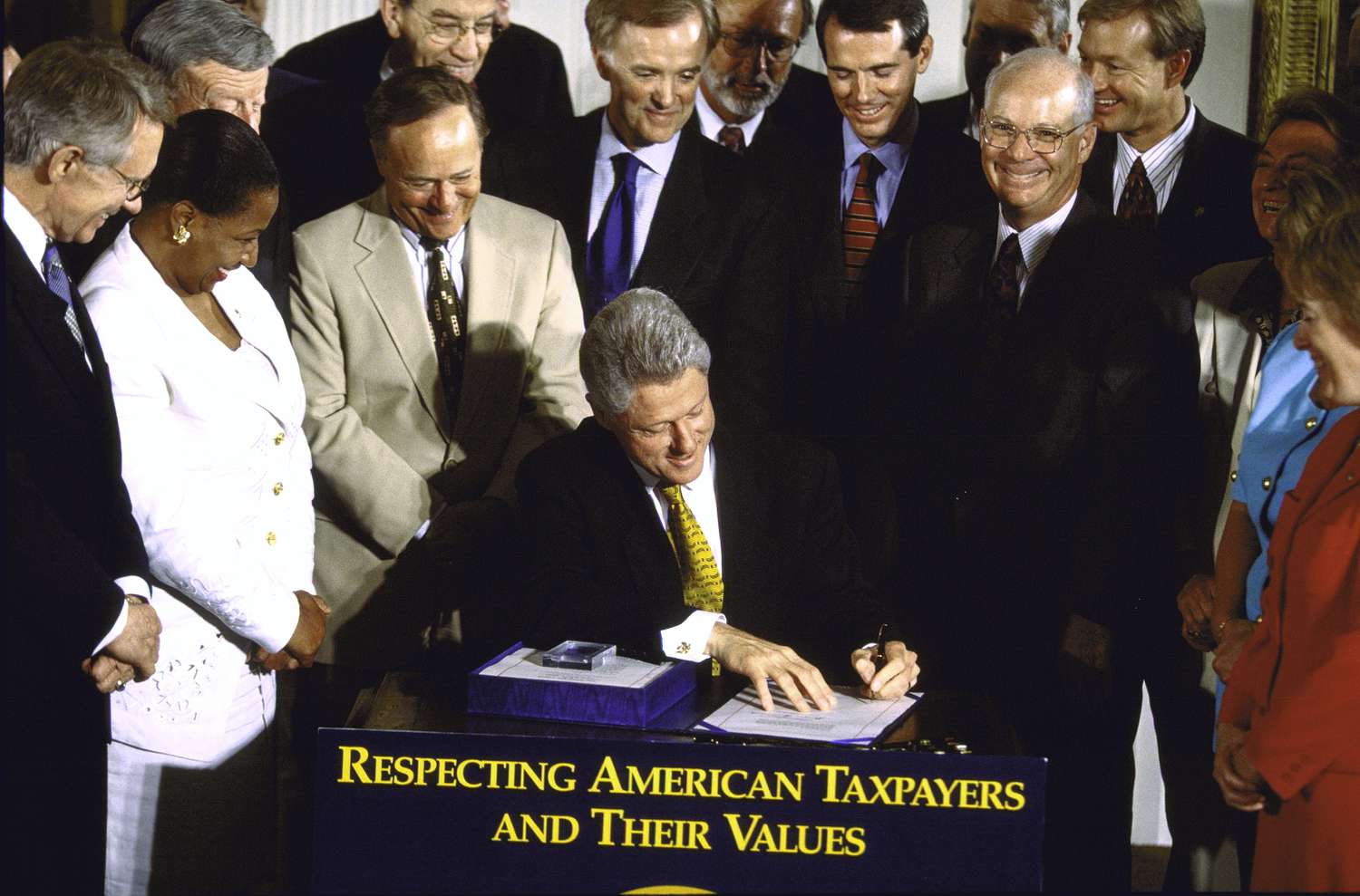 President Clinton signs a Tax Reform Act while surrounded by smiling congressional members.