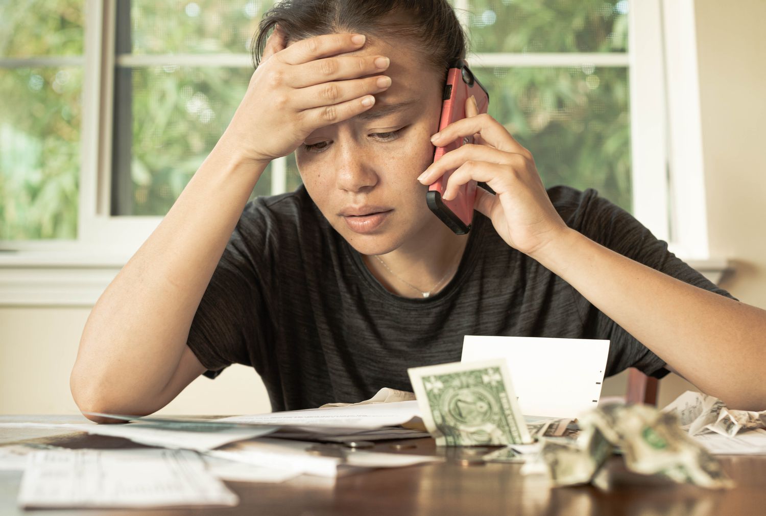 A stressed woman looks at bills on a table, holding a cell phone to her ear.