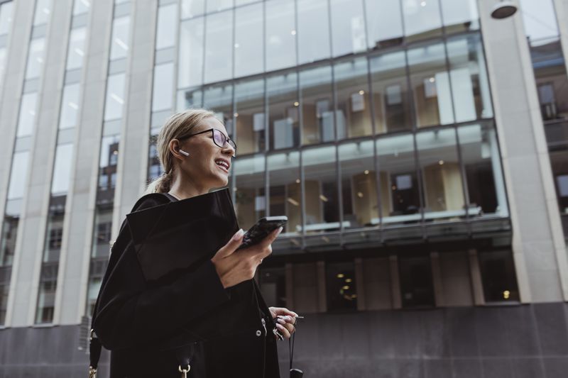 Businesswoman walking in the city surrounded by high-rises, with in-ear headphones on.