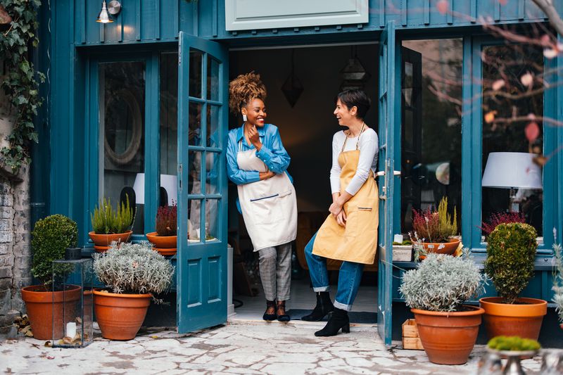 Two interior decorators and owners of a small business laugh as they share stories at the open doorway to their shop.