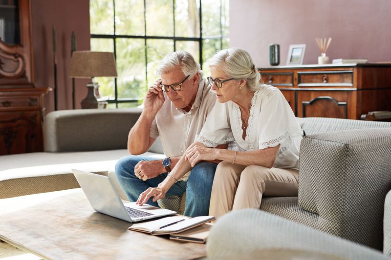 An older couple sits next to each other on a couch, looking at a laptop computer.