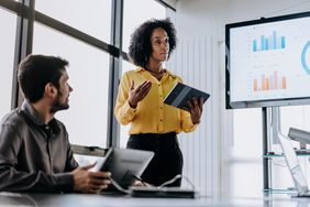 A person leads a discussion with bar charts on a screen.