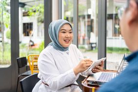 Woman sitting before a laptop holding a tablet and talking to a man sitting beside her. 