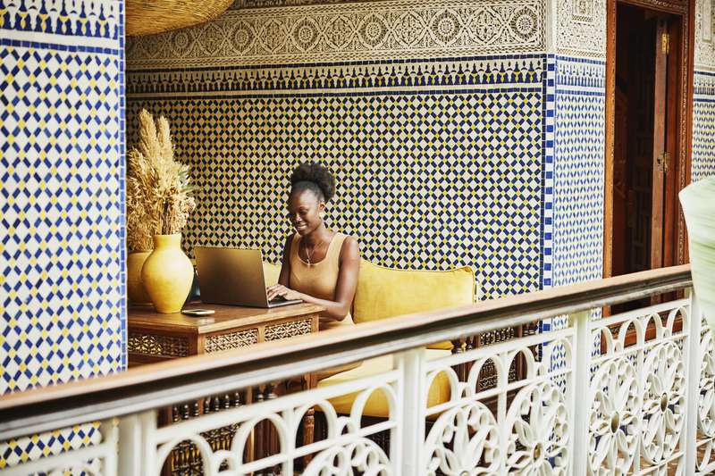 Wide shot of smiling woman working on laptop in alcove of ornately decorated riad while on vacation in Marrakech