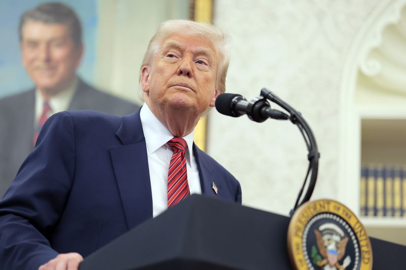 President Donald Trump stand behind a podium wearing a navy suit and a red striped tie in the oval office. 