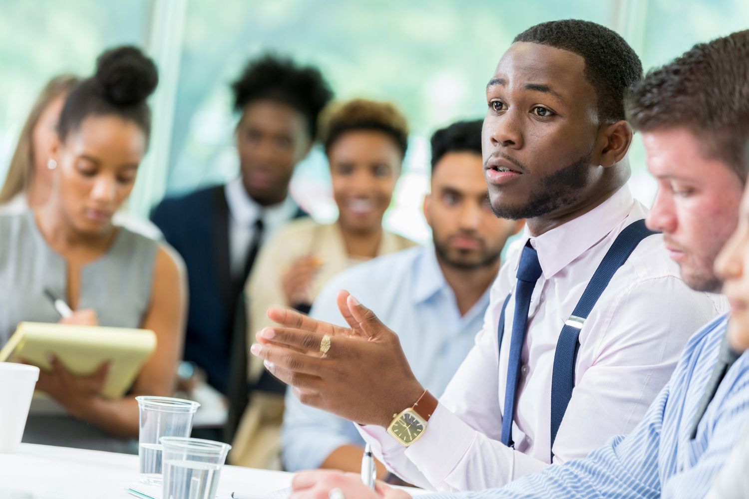 A student talks with his hands as he sits at a table in a lecture hall and poses a question to an unseen professor.