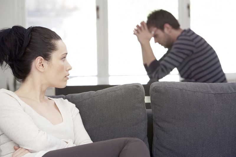 Couple looking distressed while woman sits on a couch and man holds his hands over his head.
