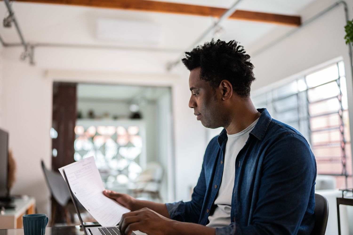 A man in a blue button down shirt sits at a table at home looking at a laptop and holding a piece of paper in his right hand