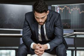 A man in a business suit sitting with his head down, stock market monitors in the background