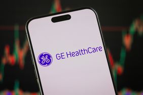 The purple GE HealthCare logo on a smartphone screen in front of a stock chart background