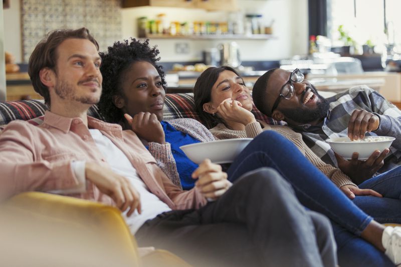 Four people sitting on a couch eating popcorn and watching tv.
