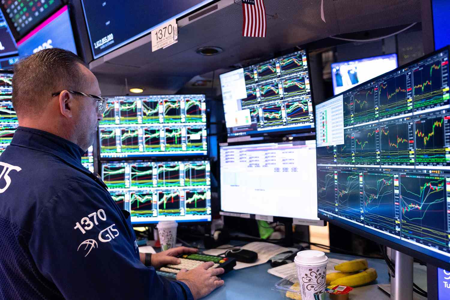 A stock trader stands at a desk looking at several computer monitors with stock charts