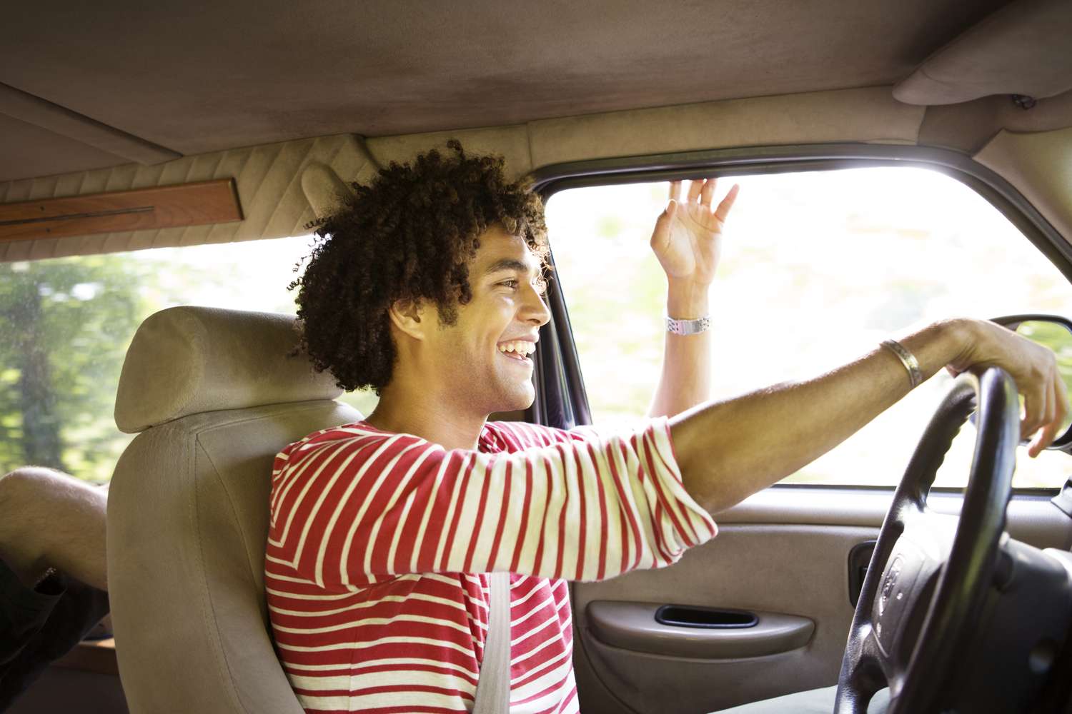 A happy, smiling man drives in a car with his hand out the window.