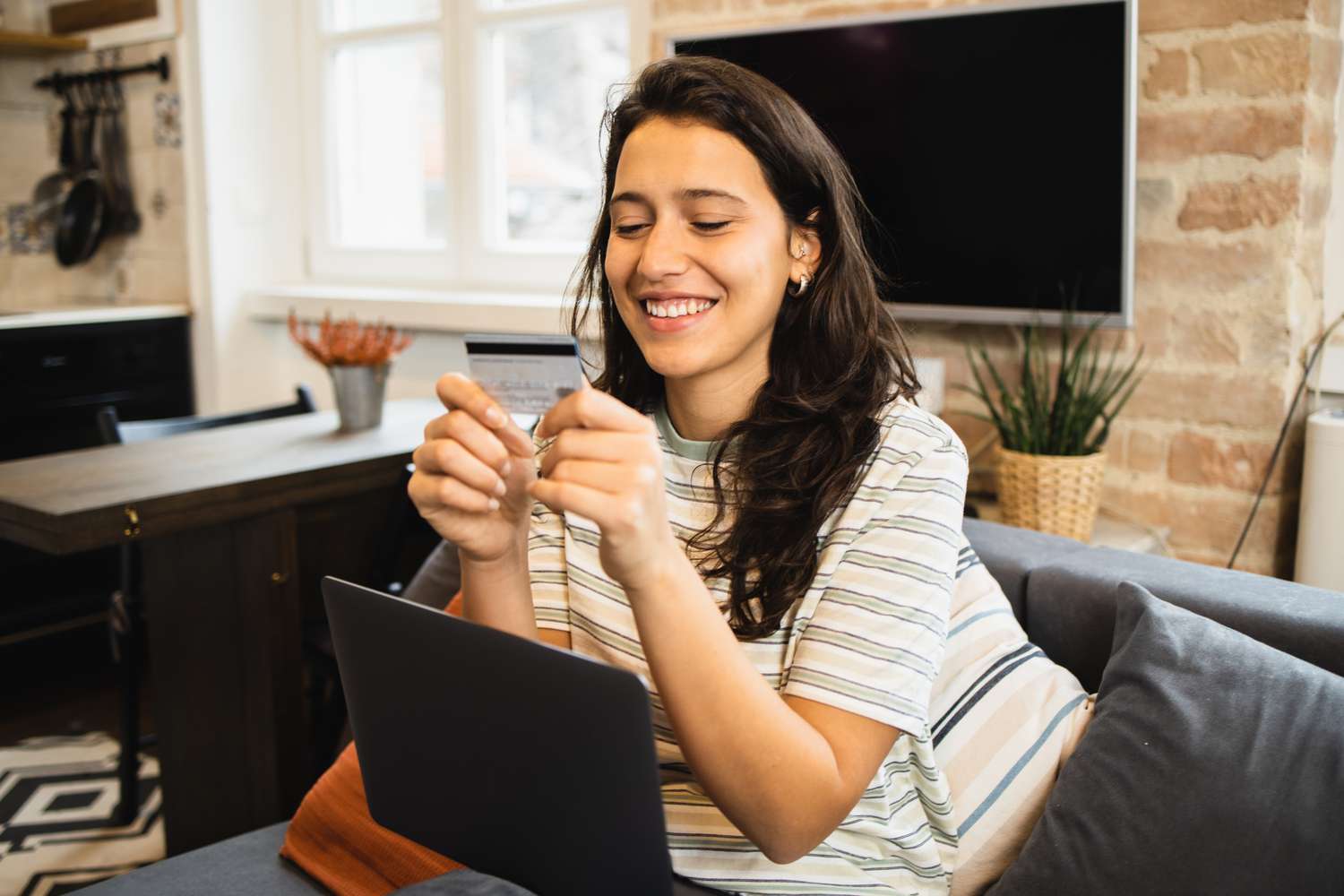 A happy young woman sits on a couch with a laptop, looking at a bank card.