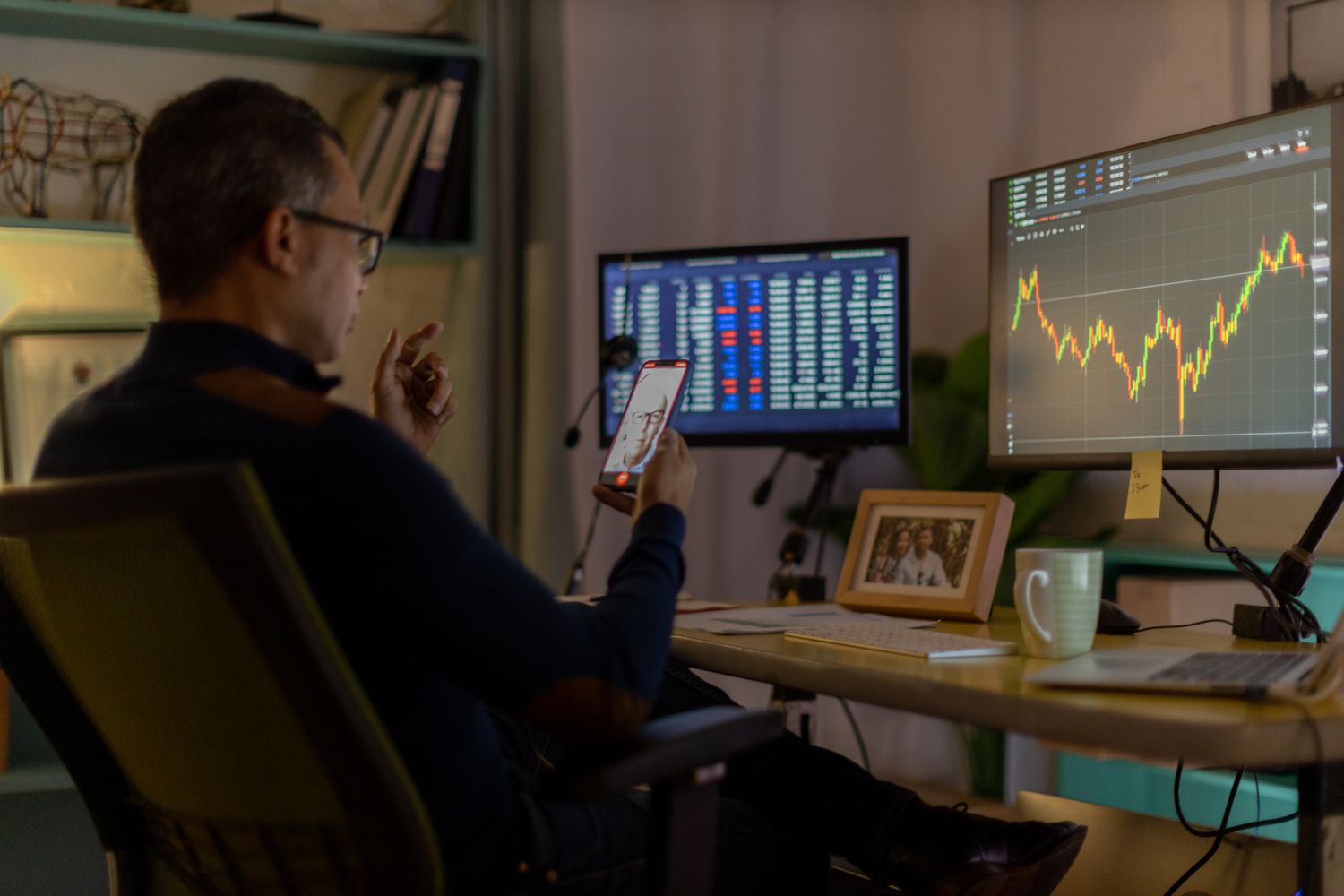 Person analyzing financial data on computer monitors in an office setting while using a smartphone