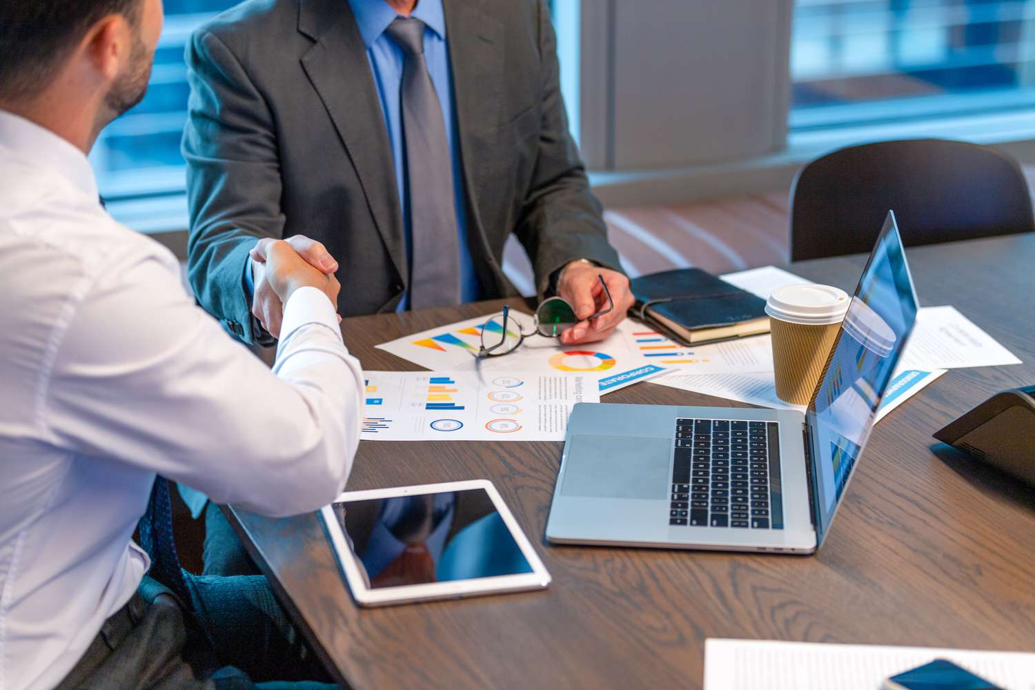 An advisor and client shaking hands while sitting at a table with laptop and tablet on the table.