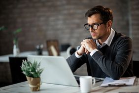 Man in his 30s sitting at a table and looking with concern at something on his laptop