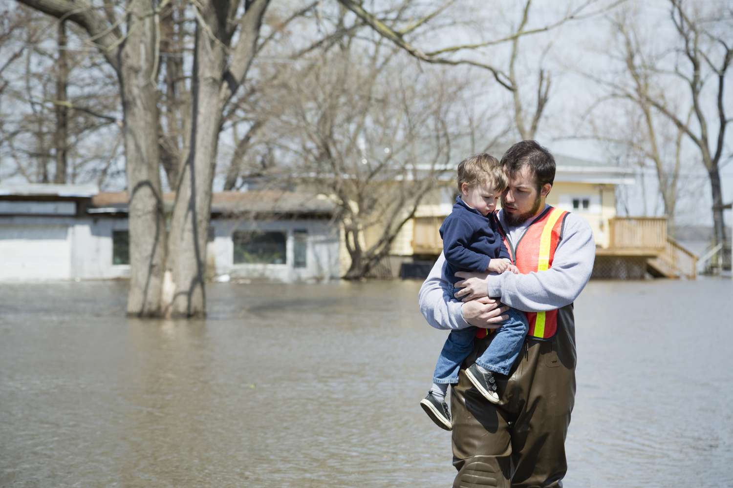 A father and son stand in a flooded street in front of a partially submerged suburban home.