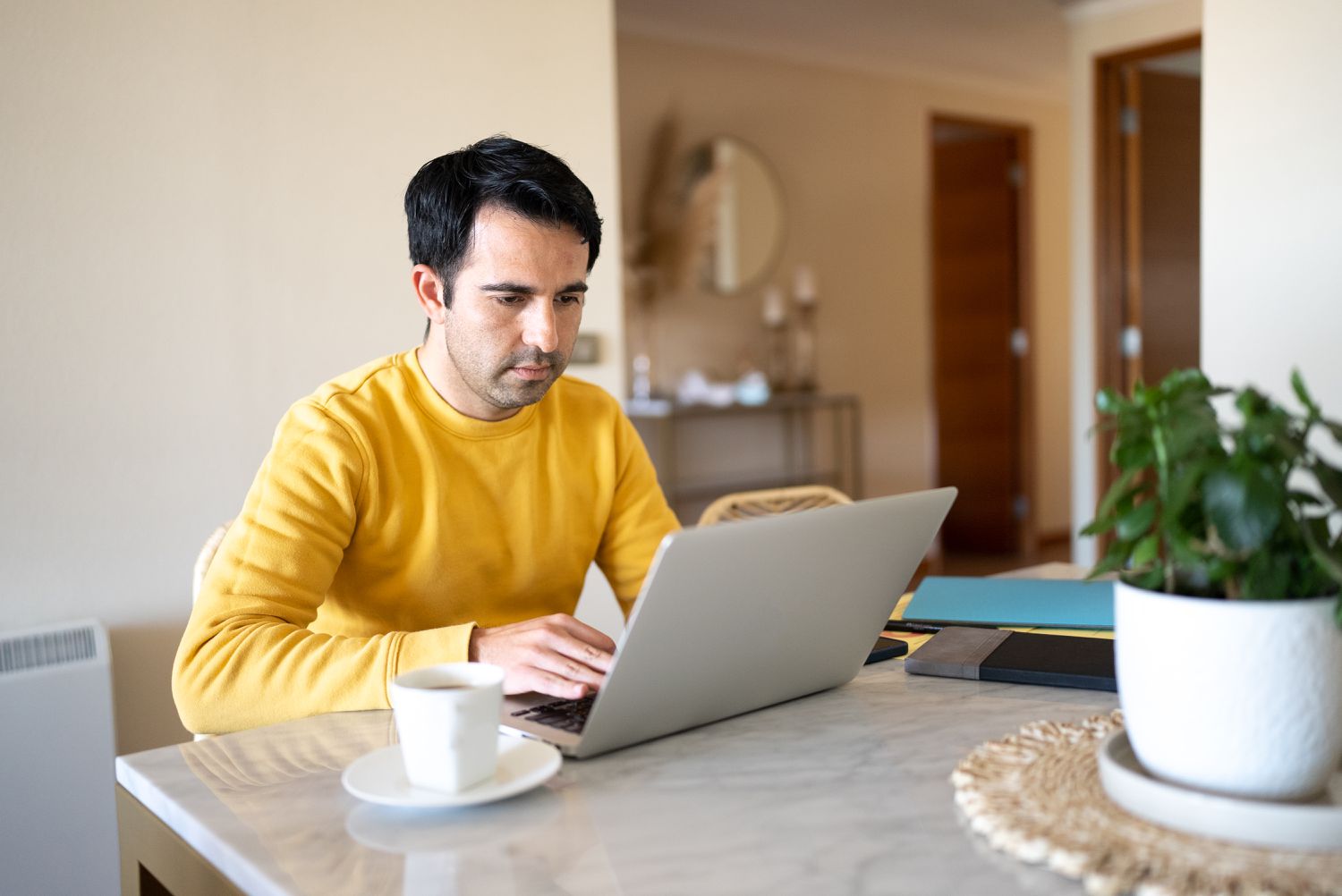A man in a yellow sweater sits at at his home desk with a laptop and a mug 