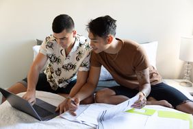 Two people sitting on a bed, looking at a laptop, with papers spread in front of them.