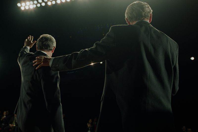 Two people in suits, with one waving to a ground from a stage in a darkened auditorium.