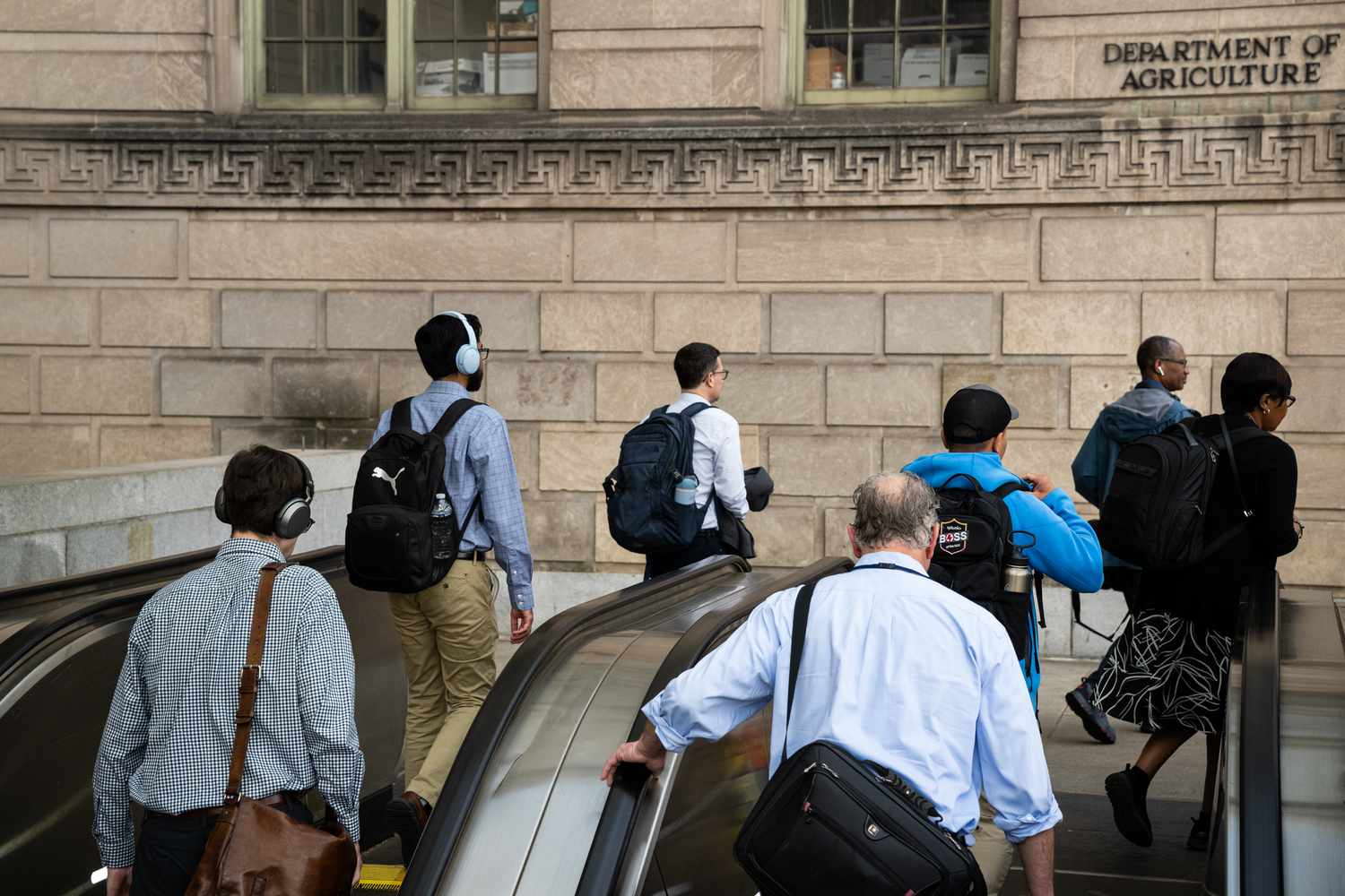 People on an escalator wear headphones and work bags outside of an ornate brick building.