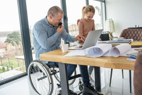 A man sitting in a wheelchair at a desk talking on the phone wit a young girl standing beside him.