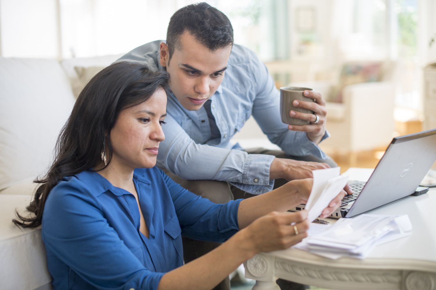 A man and a woman both wearing blue shirts sit at a coffee table with papers and a laptop; the man is holding a mug