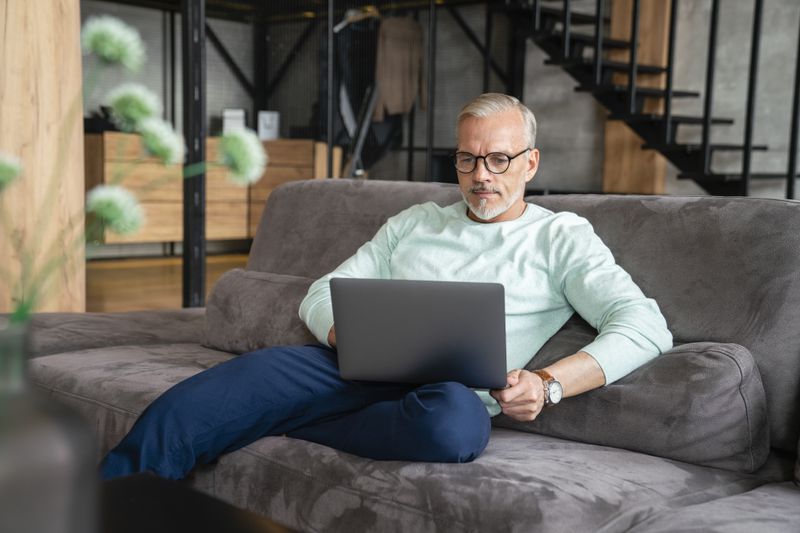 Man in in his 50s sitting on his sofa and looking intently at something on his laptop