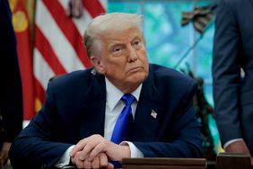 President Donald Trump sits at his desk in the oval office with his hands folded.