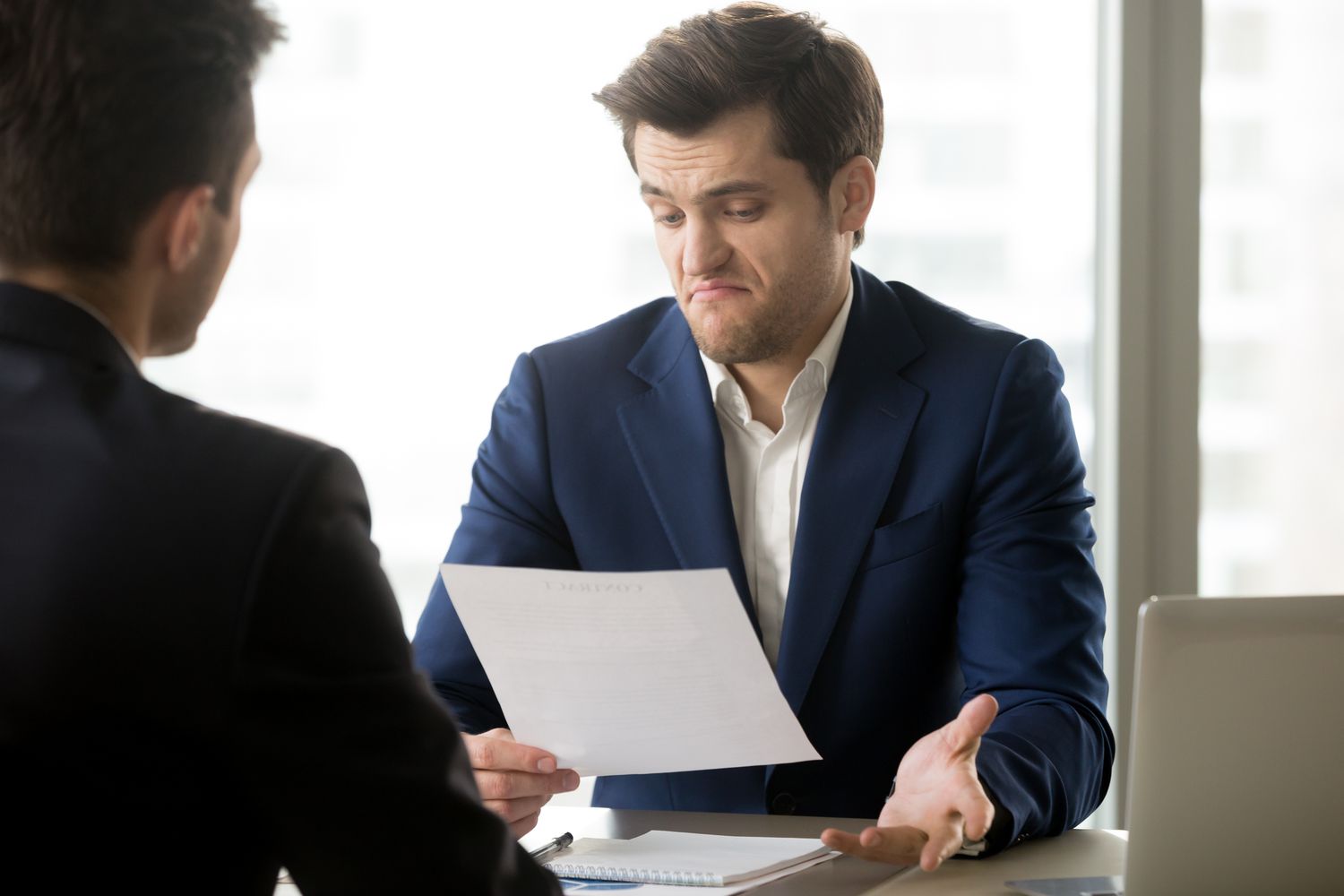 Two men talking with one shrugging while looking at a document.