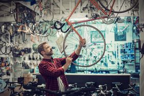 The image shows a man in a plaid red shirt inspecting a bicycle hanging from a display in a bike shop. He appears focused as he examines the wheel or frame. The shop is filled with various bicycles and bike parts hanging from the ceiling and displayed on shelves and racks in the background, creating a busy, well-stocked atmosphere. The scene conveys attention to detail and an interest in cycling.