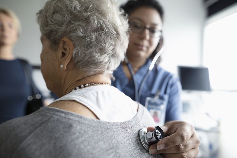 Female nurse using stethoscope on back of senior patient in clinic examination room