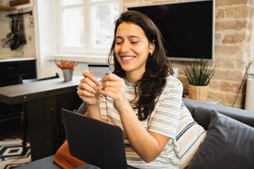 A happy young woman sits on a couch with a laptop, looking at a bank card.