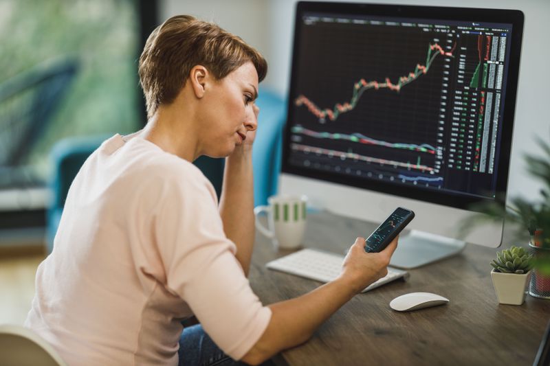 Woman at a desk, looking at a smartphone with a computer screen showing market activity in front of her
