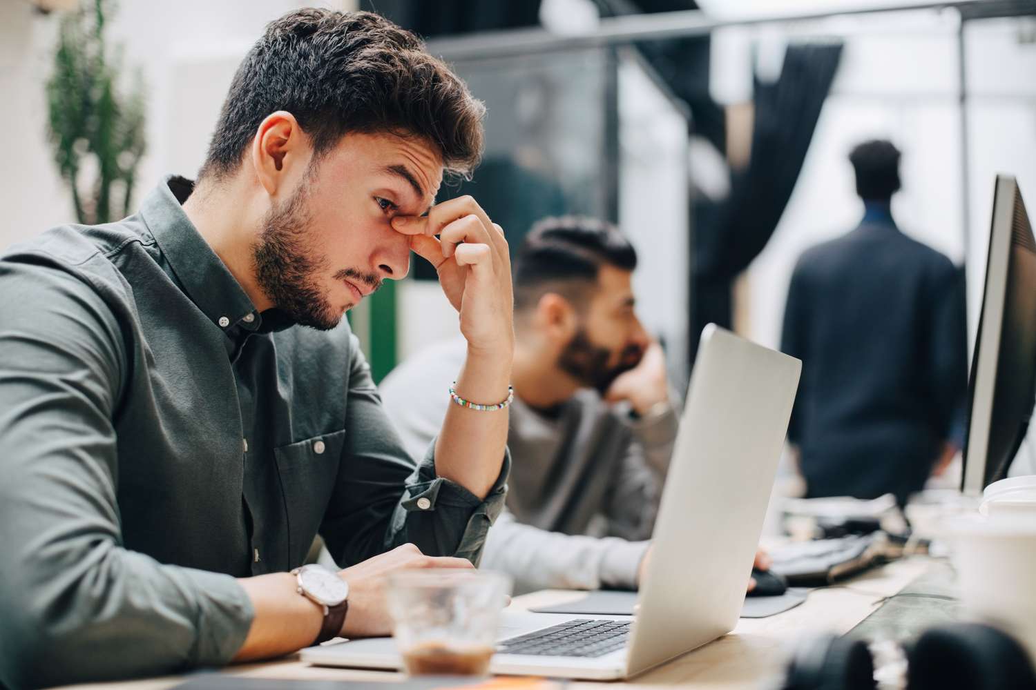 Exhausted businessman looking at laptop on desk while sitting with male coworker in office.