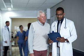 Doctor talking to an older patient while holding a clipboard and standing in a hospital corridor