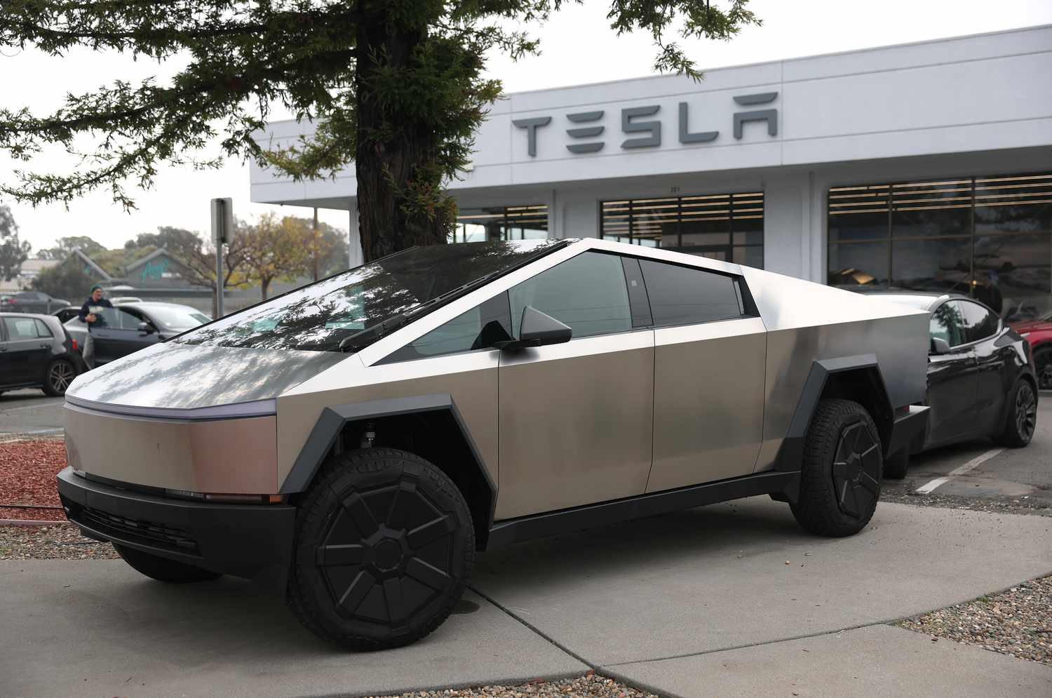 A gray Tesla Cybertruck is seen on the lot of a Tesla dealership with the office and Tesla logo in the background.