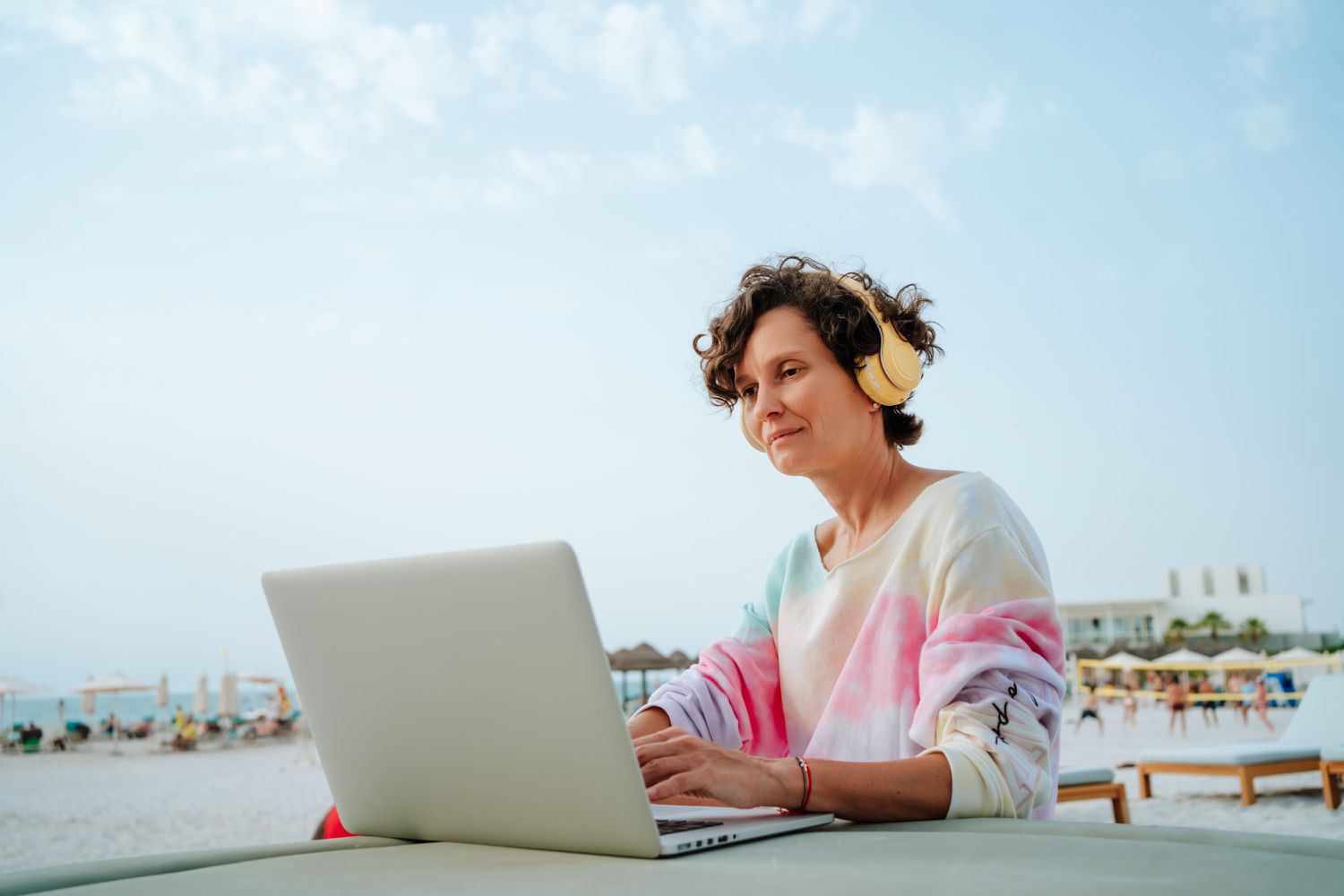 Woman sitting at a table by the beach working on a laptop with headphones on